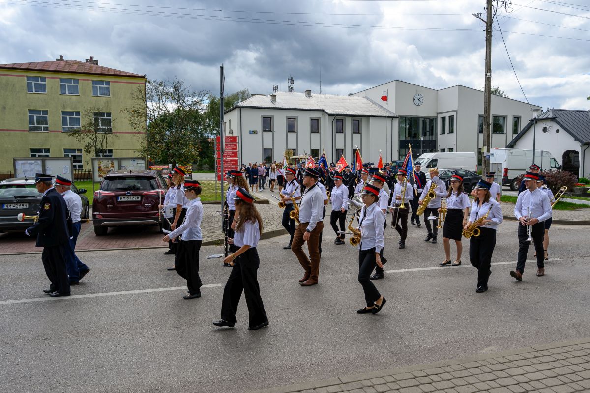 Orkiestra dęta maszeruje ulicą w czapkach i białych koszulach. W tle budynki i chmury. Flagi dodają kolorów.