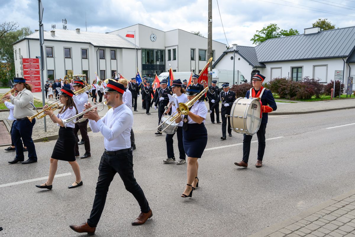 Orkiestra dęta w kolorowych mundurach maszeruje ulicą. W tle biały budynek, flaga Polski i chmury na niebie. Grają na trąbkach i bębnach.