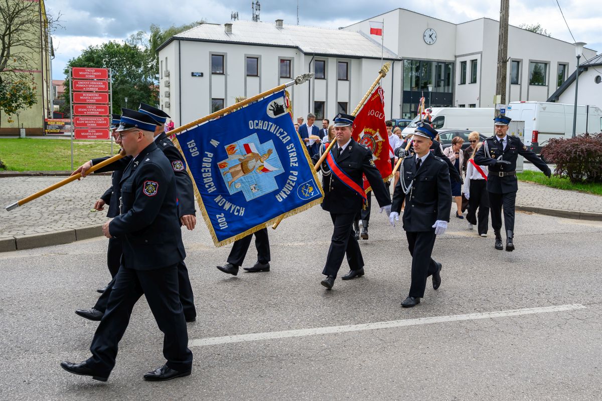 Grupa strażaków maszeruje ulicą, niosąc niebieskie i czerwone sztandary. Tło to białe budynki z flagą. Uczestnicy w ciemnych mundurach.