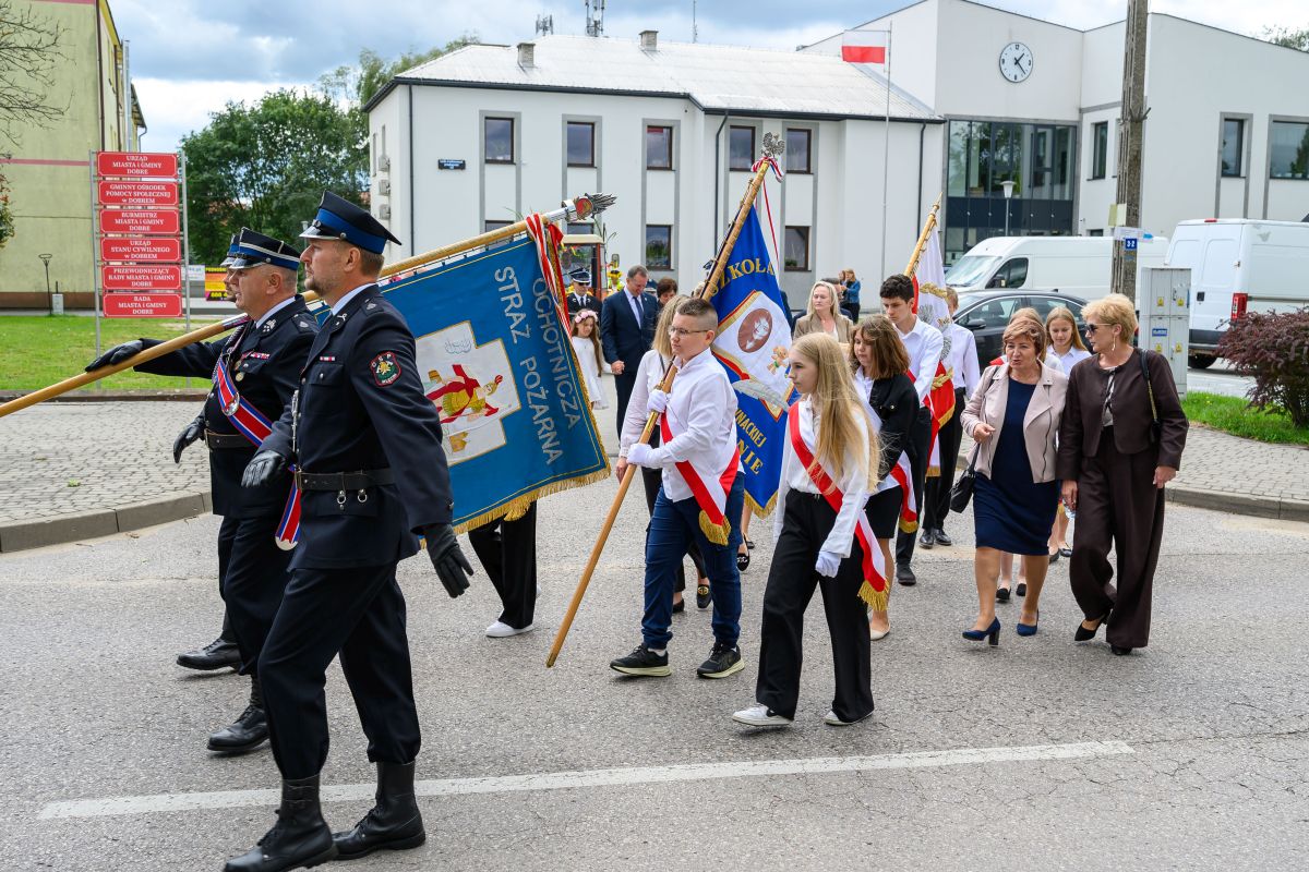 Parada na ulicy; ludzie w mundurach i młodzież z flagami. Tło z budynkami i drzewami, niebo pochmurne. Kolory jasne i kontrastowe.