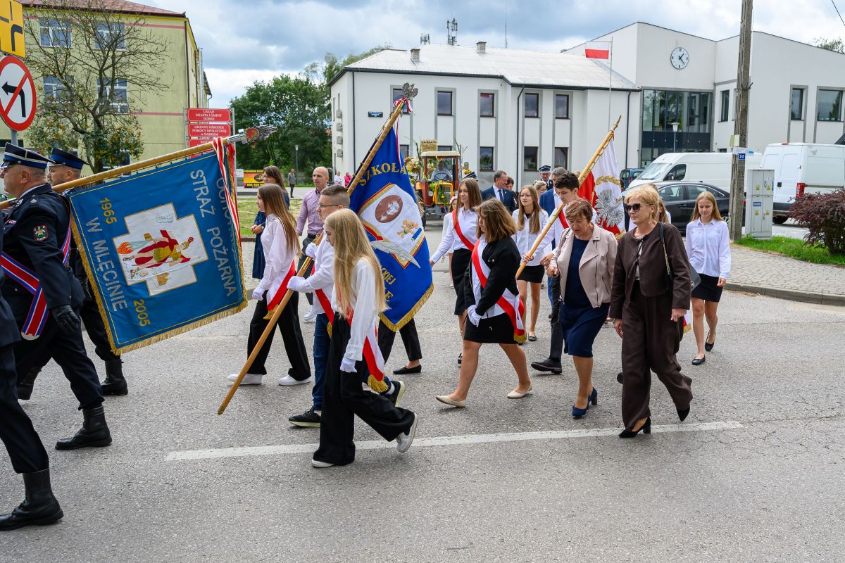 Grupa młodych ludzi maszeruje z niebieskimi i białymi chorągwiami na ulicy. Ubrani są w białe koszule z czerwonymi szarfami. W tle znajduje się budynek i zielone drzewa.