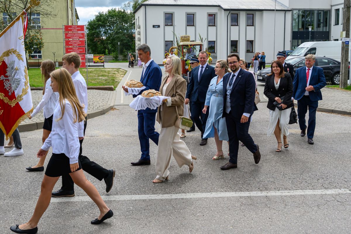 Grupa ludzi w eleganckich ubraniach idzie ulicą przy budynkach, trzymając chleb na białych serwetkach. Flaga z orłem i czerwonym tłem. Otoczenie jest zielone z chmurami na niebie.