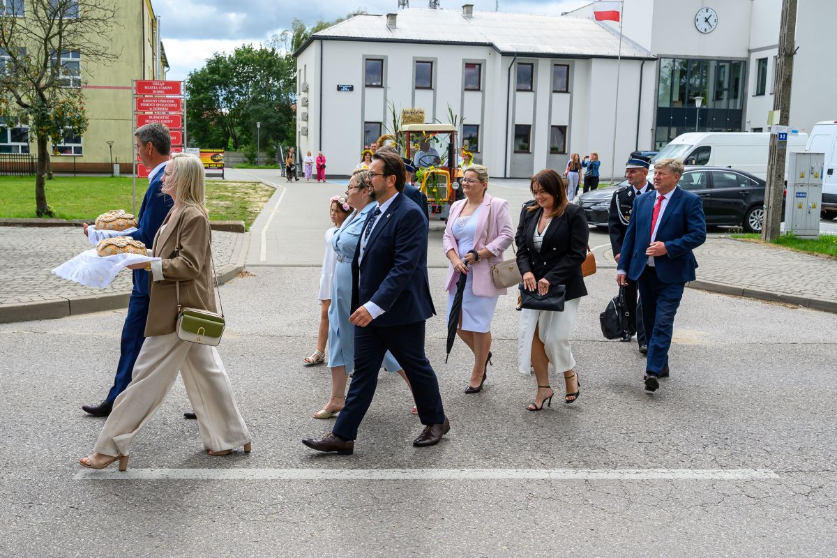 Grupa ludzi w formalnych strojach idzie ulicą, dwoje niesie chleb. Tło: biały budynek i szare niebo. Obok powiewa polska flaga.