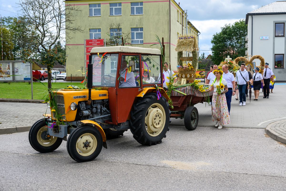 Na zdjęciu widoczny jest kolorowy traktor z przyczepą ozdobioną plonami i słomianymi wieńcami. W tle stoi żółto-zielony budynek, a na ulicy widoczna jest grupa osób. Sceneria przypomina tradycyjny festiwal dożynkowy.