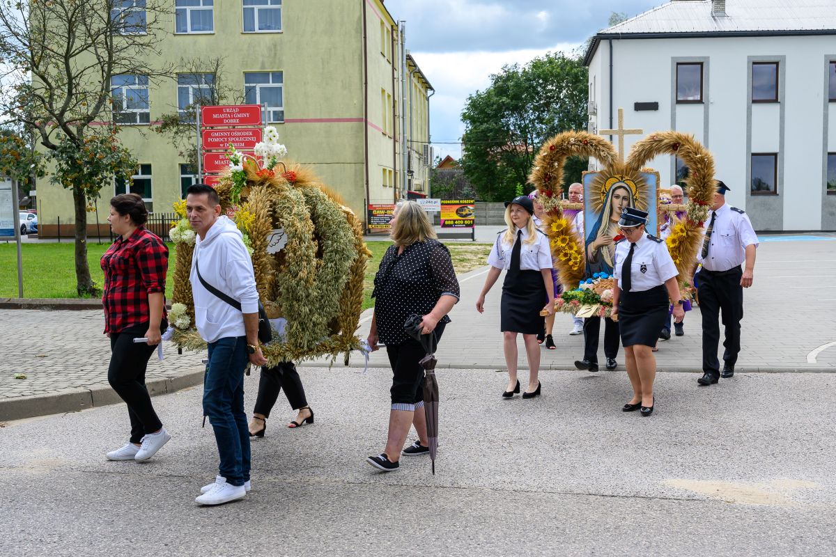 Grupa ludzi niesie ozdobne wieńce z zboża, na jednym obraz Matki Boskiej. Tło to budynki i drzewa. Atmosfera jest uroczysta.