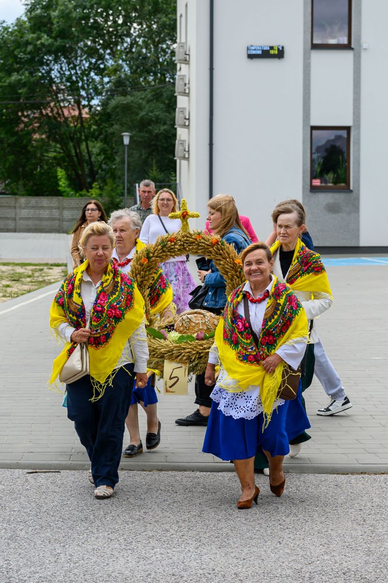 Grupa kobiet w tradycyjnych, kolorowych strojach niesie dekoracyjny wieniec. Tło to budynek i drzewa. Atmosfera świąteczna.