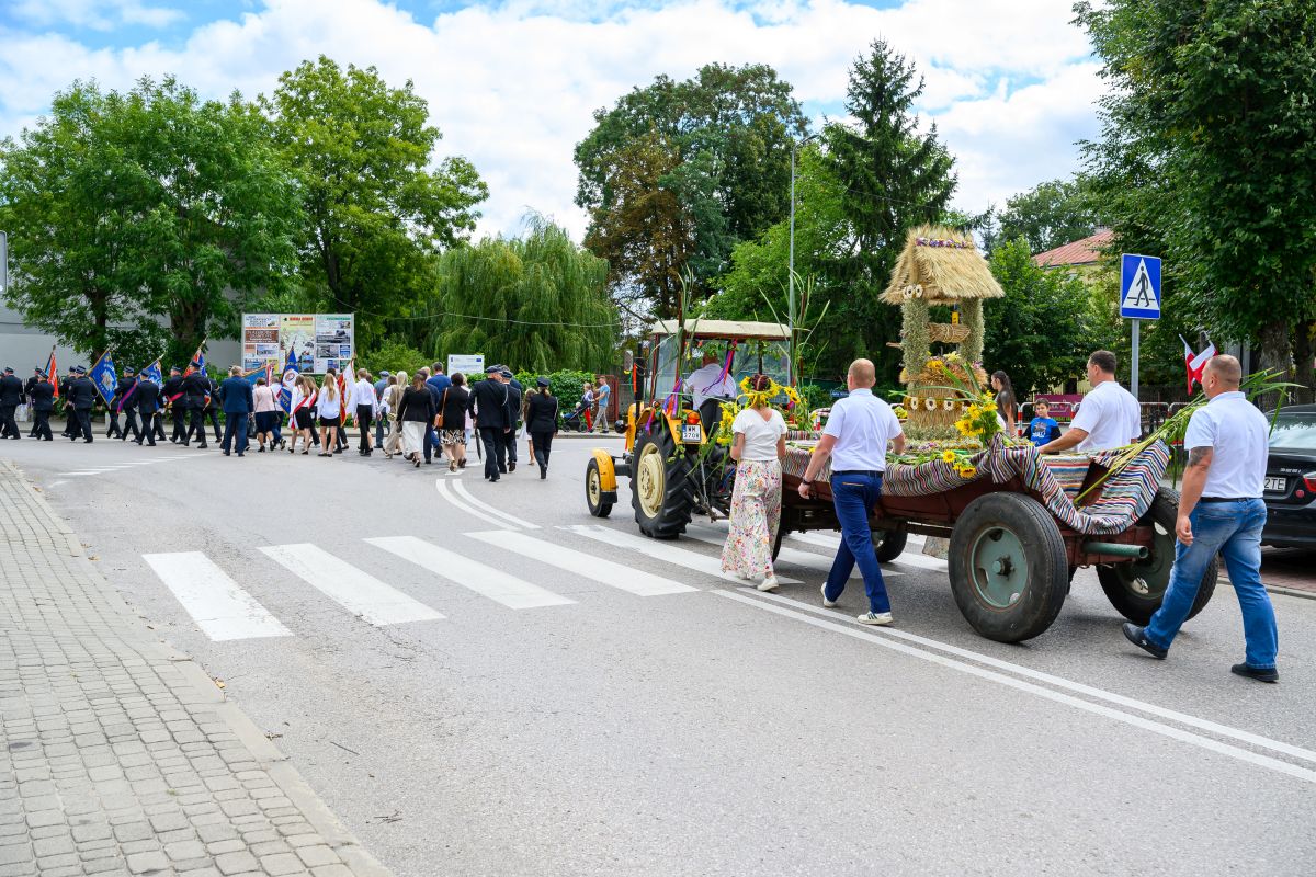 Parada na ulicy: traktor ciągnący kolorowo udekorowaną przyczepę. W tle uczestnicy niosą flagi. Zielona sceneria dookoła.