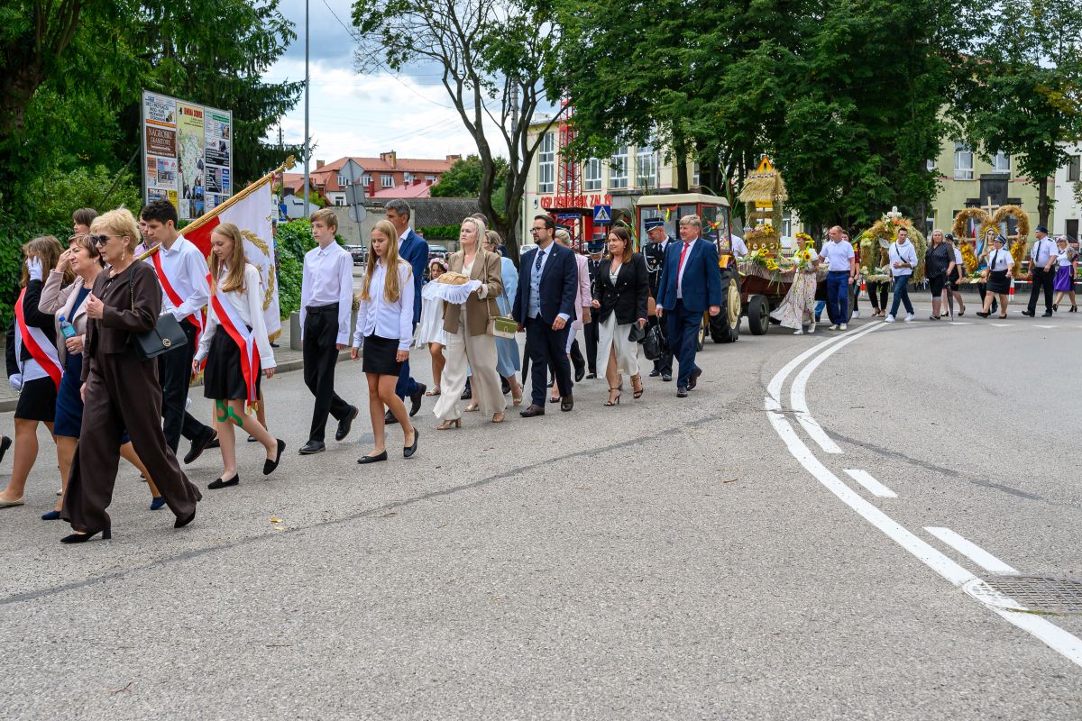 Uroczysta procesja idzie ulicą, ludzie ubrani formalnie, wśród zieleni i miejskich budynków. Tło stanowią drzewa i budynki. Kolory dominujące to zieleń, biel oraz czerwony akcent flag.