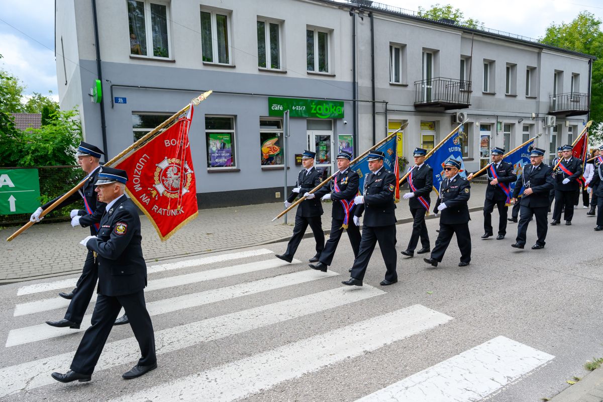 Grupa strażaków w mundurach maszeruje z kolorowymi sztandarami na przejściu dla pieszych. Budynki w tle są szare, a pogoda pochmurna. Na banerach dominuje kolor czerwony i niebieski.