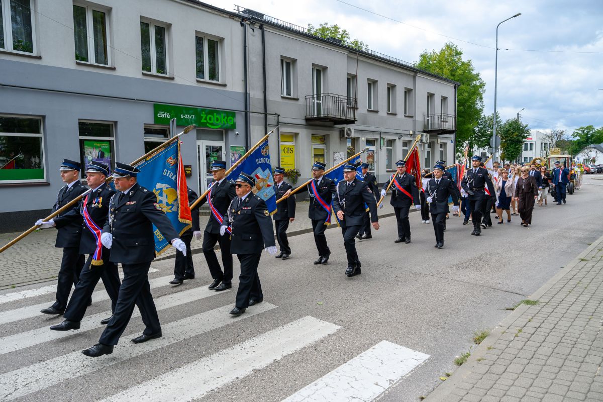 Parada strażaków w mundurach maszerujących ulicą z flagami. Tło stanowią budynki i drzewa. Kolory to głównie niebieski i czerwony.