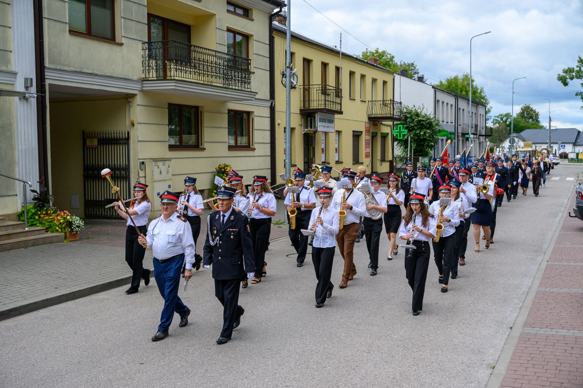 Orkiestra maszeruje ulicą w kolorowych mundurach, grając instrumenty. Tło stanowią budynki i drzewka. Po prawej stronie jest chodnik z kostki.