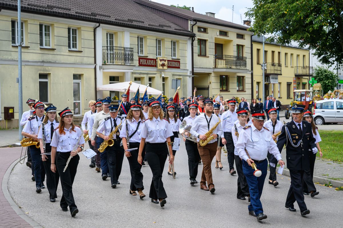 Orkiestra dęta maszeruje ulicą, muzycy w białych koszulach i czapkach. W tle są kremowe budynki. Zieleń po prawej stronie.