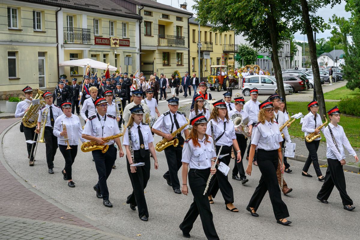 Parada orkiestry dętej w białych koszulach i czapkach na ulicy. Tło to budynki z szyldami i zielone drzewa. Zanurzeni w muzyce, maszerują.