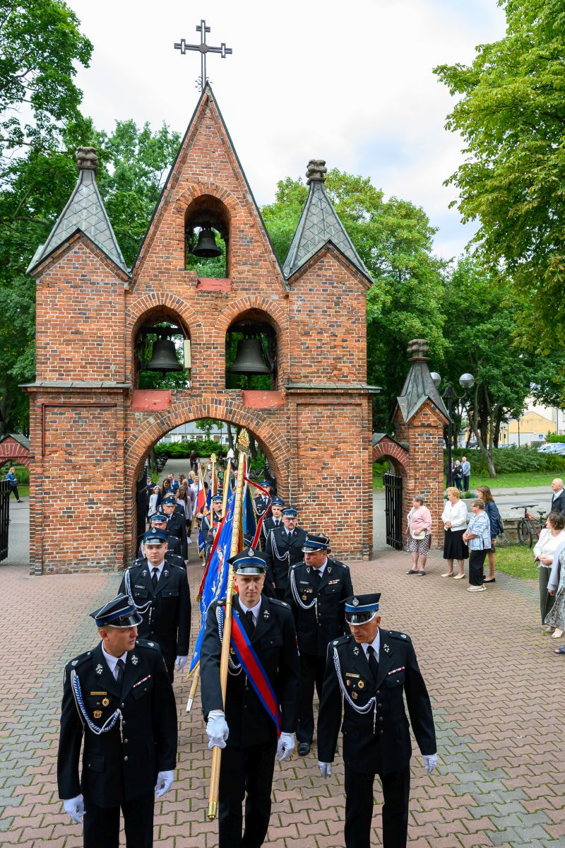 Parada strażaków w czarnych mundurach przed ceglaną dzwonnicą. Kolory to czerwień cegieł i zieleń drzew. Ludzie oglądają z boku.