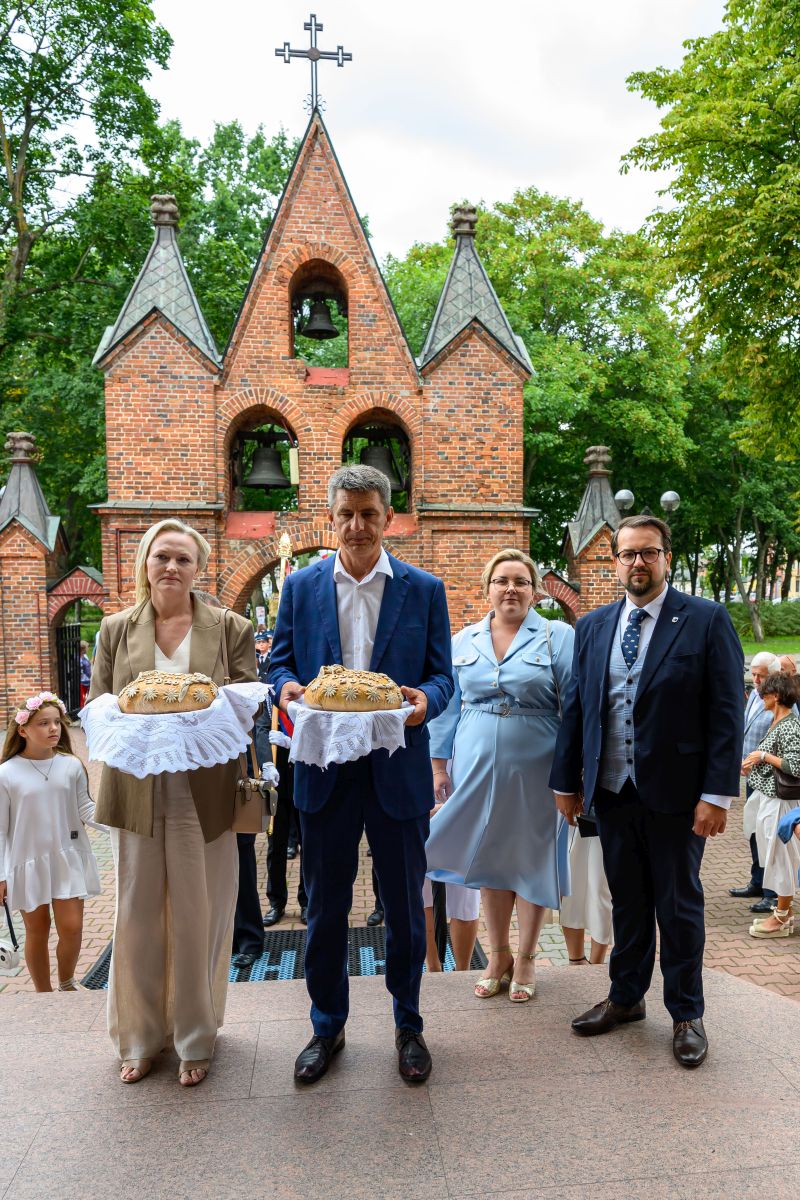 Cztery osoby w eleganckich strojach trzymają chleby na białych serwetkach przed ceglanym budynkiem z dzwonami. W tle zielone drzewa i krzyż na szczycie. Obecni są również ludzie w tle, co wskazuje na uroczystość.