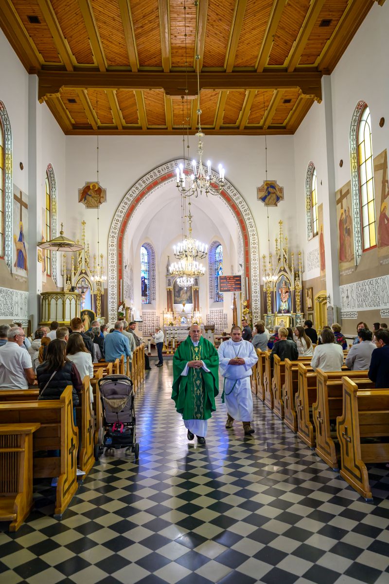 Wnętrze kościoła z drewnianym sufitem i żyrandolami. Ksiądz w zielonej szacie przechodzi przez nawy pełną ludzi. Tło zdobią witraże i obrazy religijne.