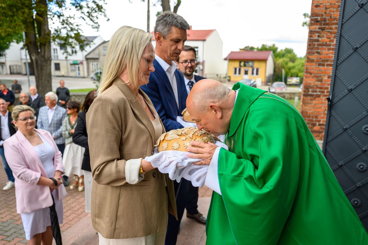 Na zdjęciu widać kobietę i mężczyznę trzymających chleb, a duchowny w zielonym stroju go całuje. W tle widoczni są ludzie, budynki i drzewa. Kolory są jasne i naturalne, a scena odbywa się na zewnątrz.