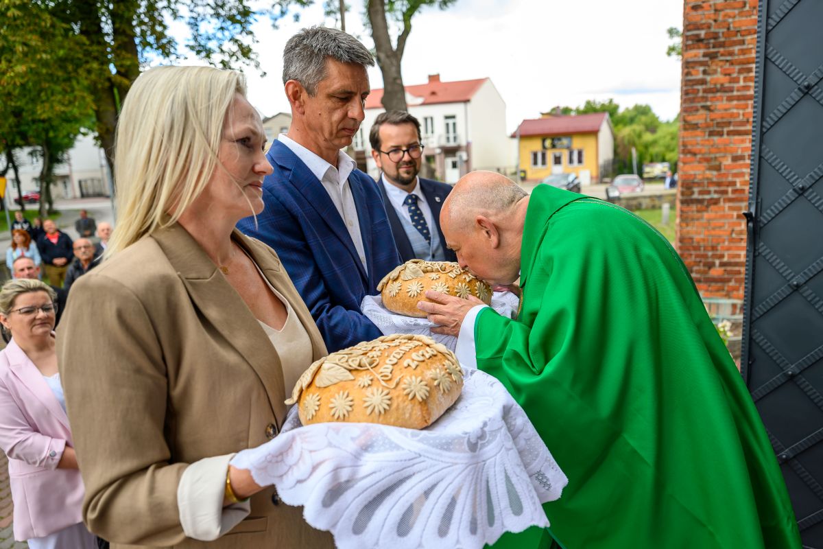Mężczyzna w zielonym ornacie pochyla się nad chlebami ozdobionymi ornamentami. Obok para trzyma chleb na białych serwetkach. Tło to budynki i drzewa.