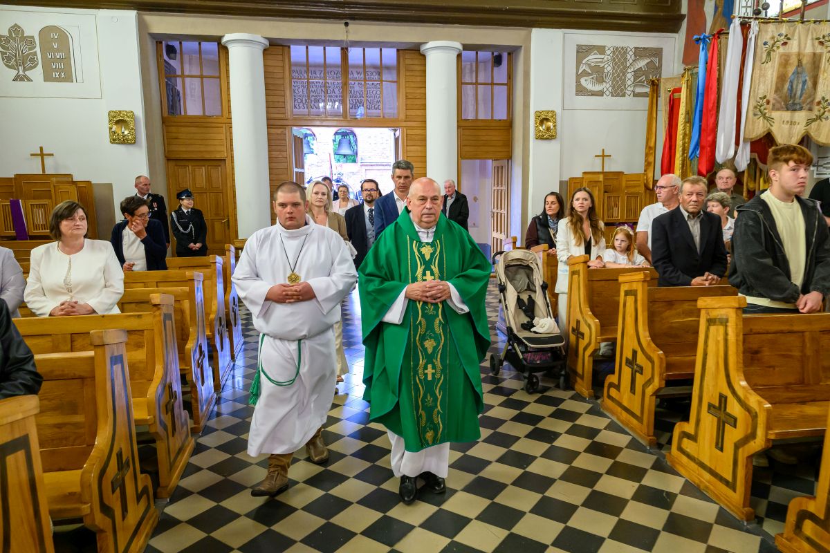 Procesja w kościele; ksiądz w zielonej szacie i ministrant w białej idą środkiem między ławkami. Drewniane ławki, kolorowa posadzka. Uczestnicy stoją po bokach, w tle witraże.