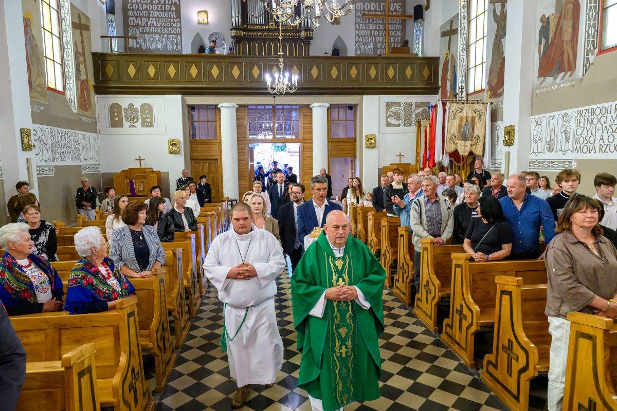 W jasnym kościele ksiądz w zielonej szacie prowadzi procesję. Na ławkach siedzą wierni w strojach ludowych. Ściany zdobią dekoracje i teksty biblijne.