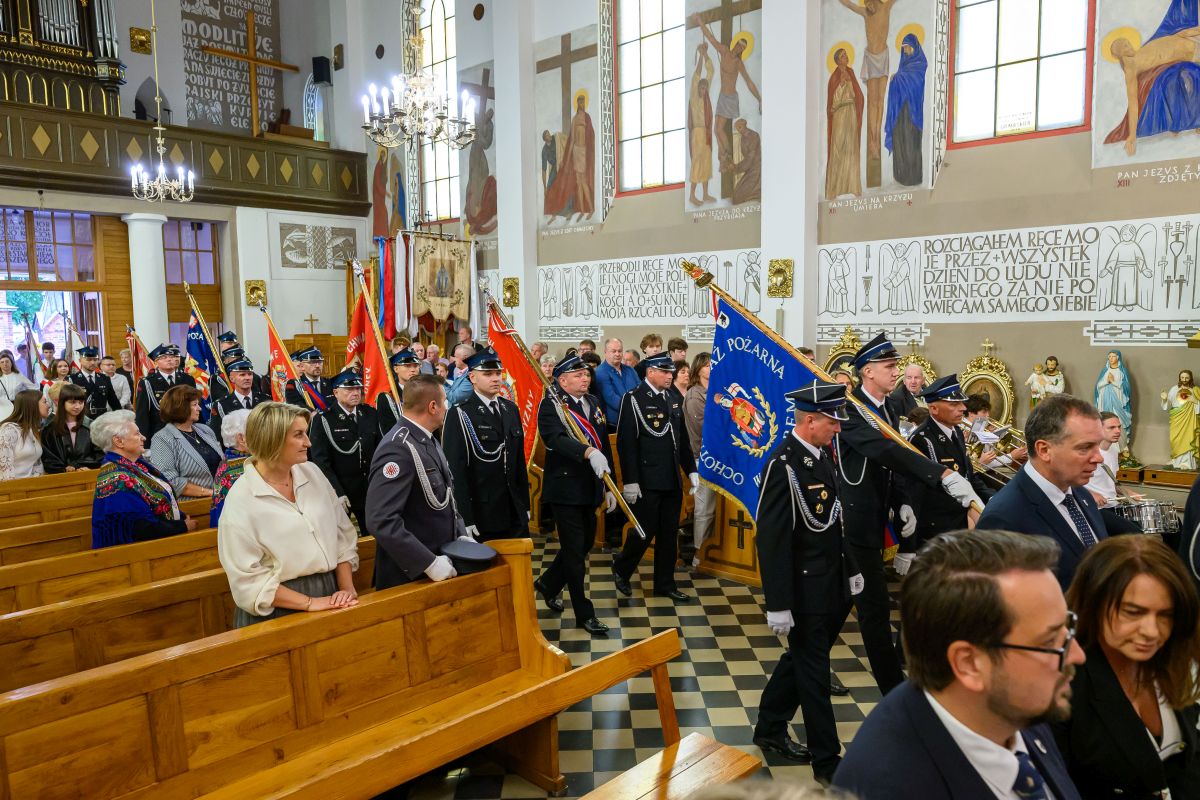 Procesja strażaków w mundurach z flagami w kościele pełnym ludzi. Ściany zdobią freski i napisy, a wnętrze jest jasne.
