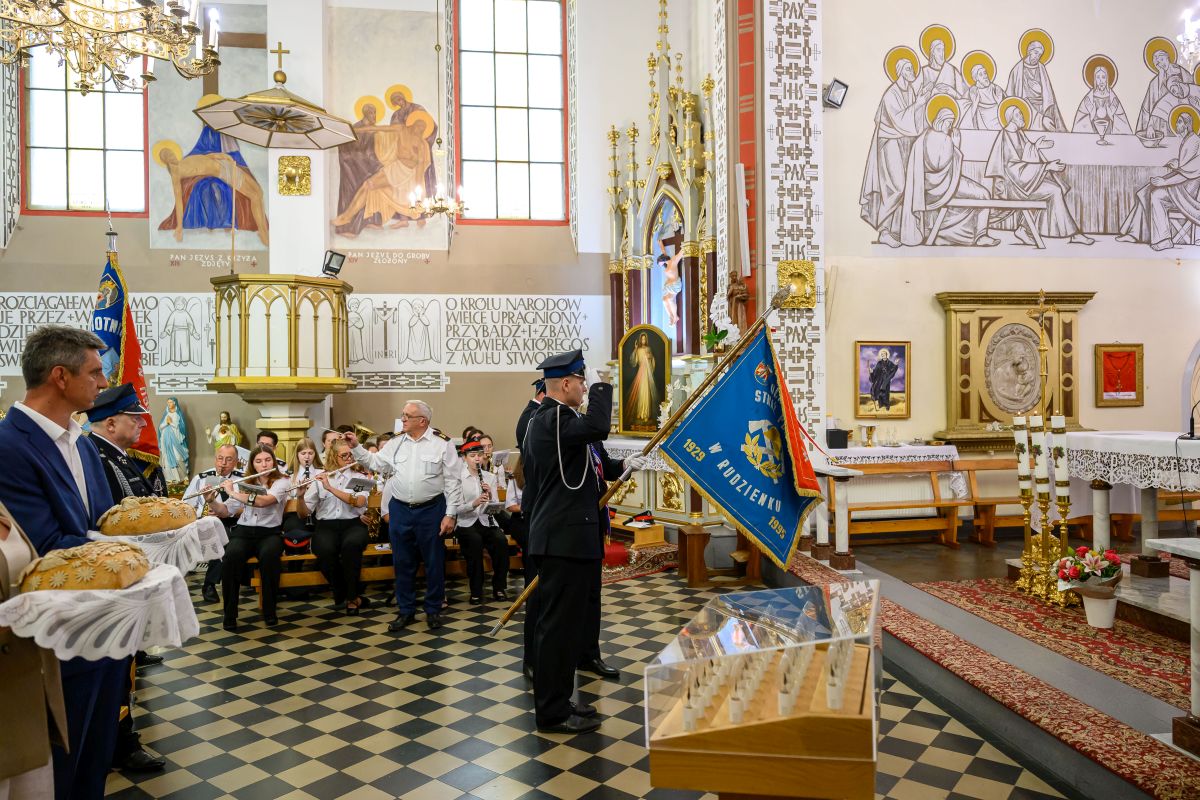 Kościół z wiernymi podczas ceremonii, orkiestra dęta na tle witraży. Dominuje kolor biały i złoty we wnętrzu. Procesja z flagą strażacką.