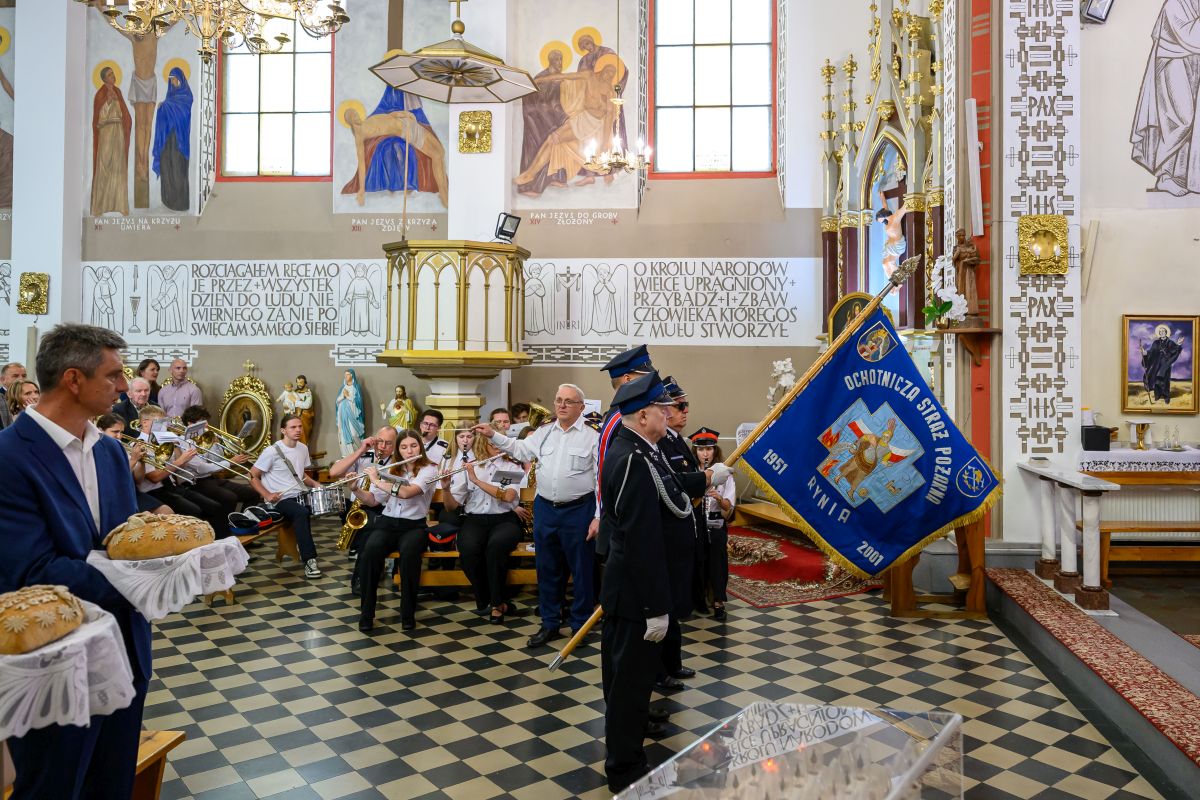 Uroczystość w jasnym kościele z orkiestrą i sztandarem straży pożarnej. Na przodzie mężczyzna trzyma chleb w białych serwetkach. Ściany zdobią religijne malowidła i złote detale.