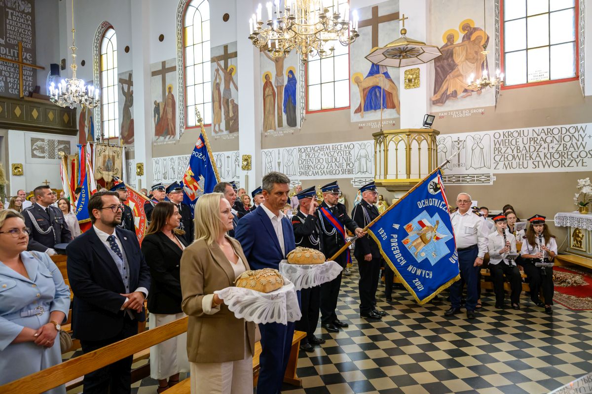 Na obrazie widoczna jest uroczystość w kościele z ludźmi niosącymi chleby. Tło zdobią kolorowe freski i flagi strażackie. Dominują kolory biały, niebieski i złoty.
