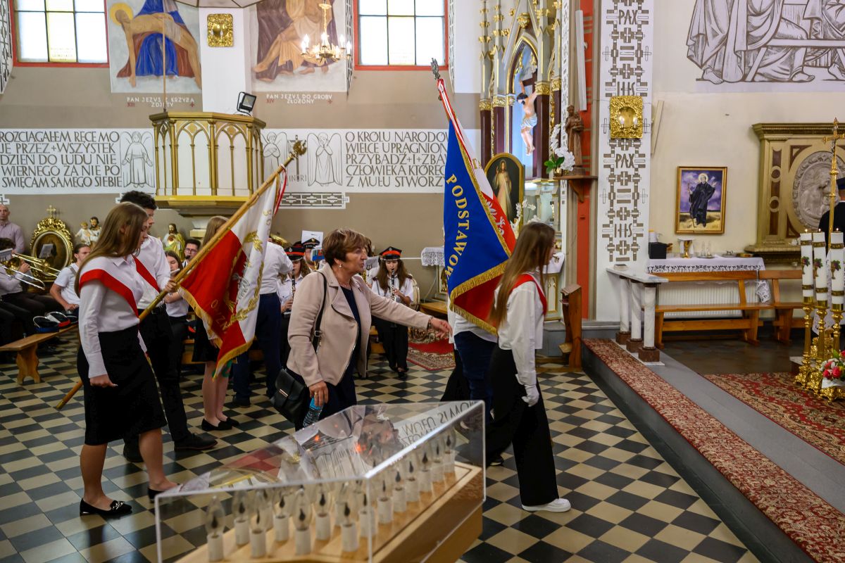 Grupa uczniów z flagami wchodzi do kościoła. Tło ozdobione malowidłami i złotymi elementami. Podłoga w czarno-białą szachownicę.