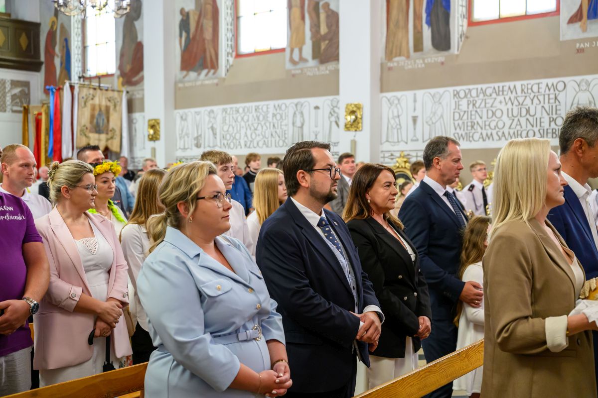 Zdjęcie przedstawia grupę ludzi w kościele podczas nabożeństwa. Widać kolorowe witraże i malowidła ścienne w tle. Osoby są elegancko ubrane i stoją w rzędzie.