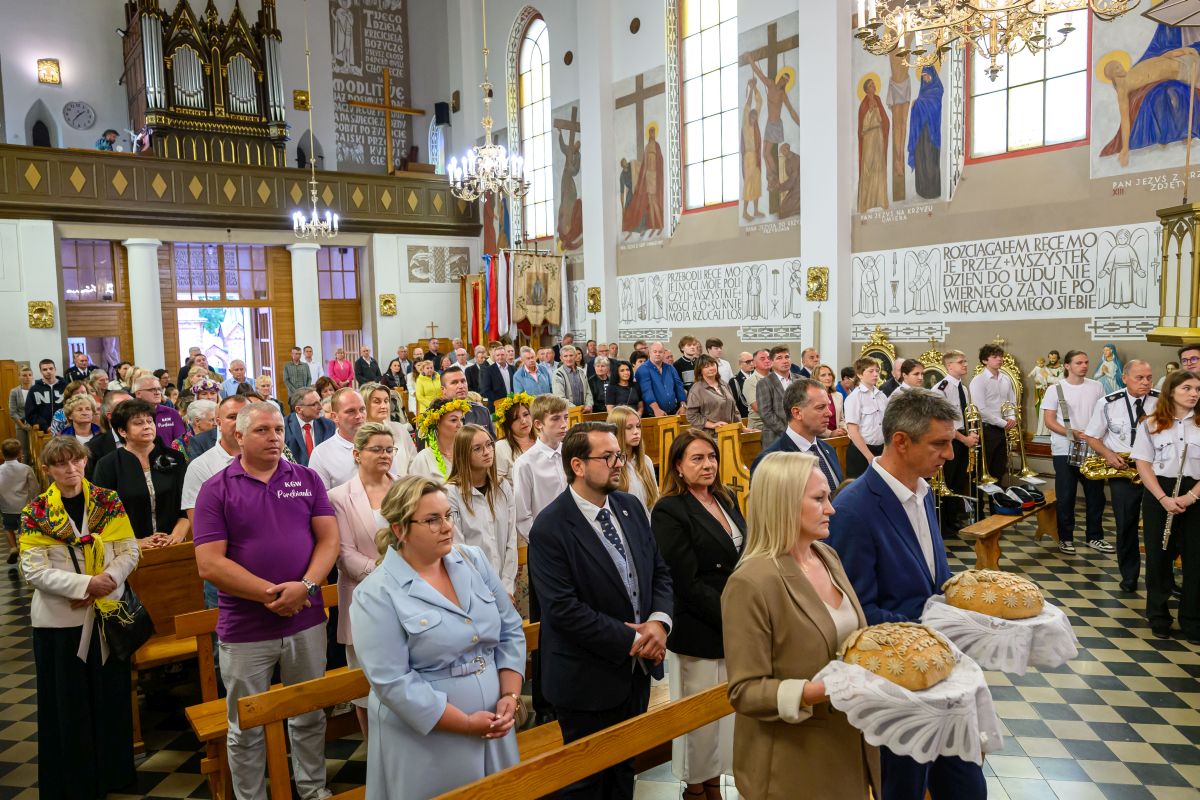 Wnętrze kościoła pełne ludzi uczestniczących w uroczystości. Dominują kolory białe i brązowe, mury zdobią barwne freski. Na pierwszym planie para trzyma ozdobne chleby.