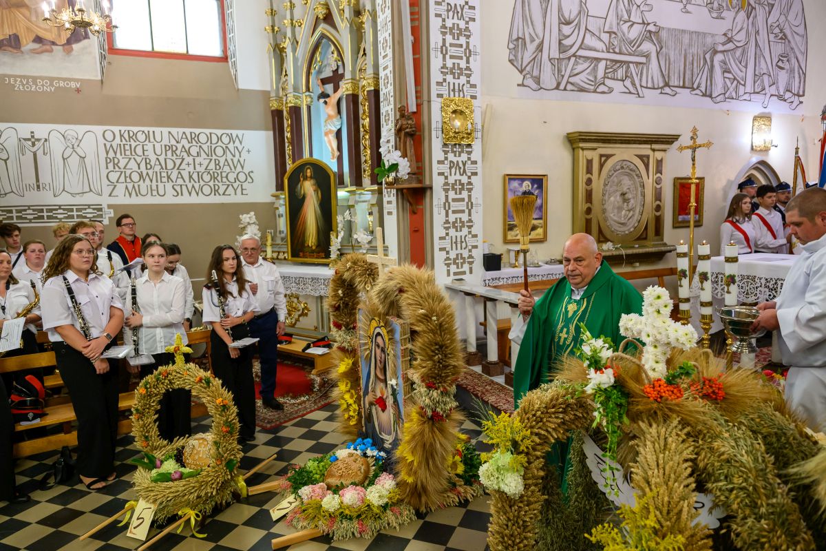 Ceremonia kościelna z kapłanem w zielonym ornacie. Otoczenie ozdobione wieńcami z kłosków i kwiatów. W tle biało-złote elementy ołtarza.