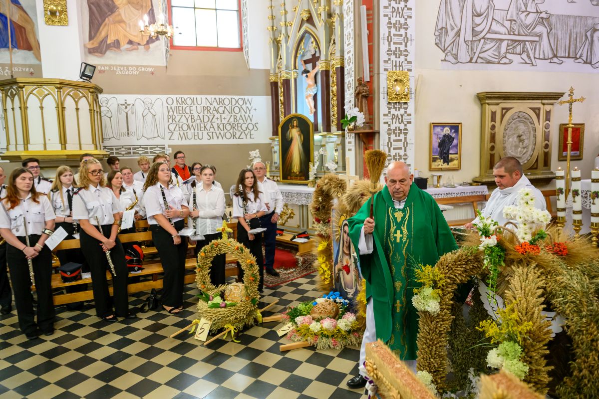 Kapłan w zielonym ornacie błogosławi wieńce dożynkowe w kościele. Obok grupa osób w białych koszulach. Tło ozdobione jest religijnymi obrazami i rzeźbami.