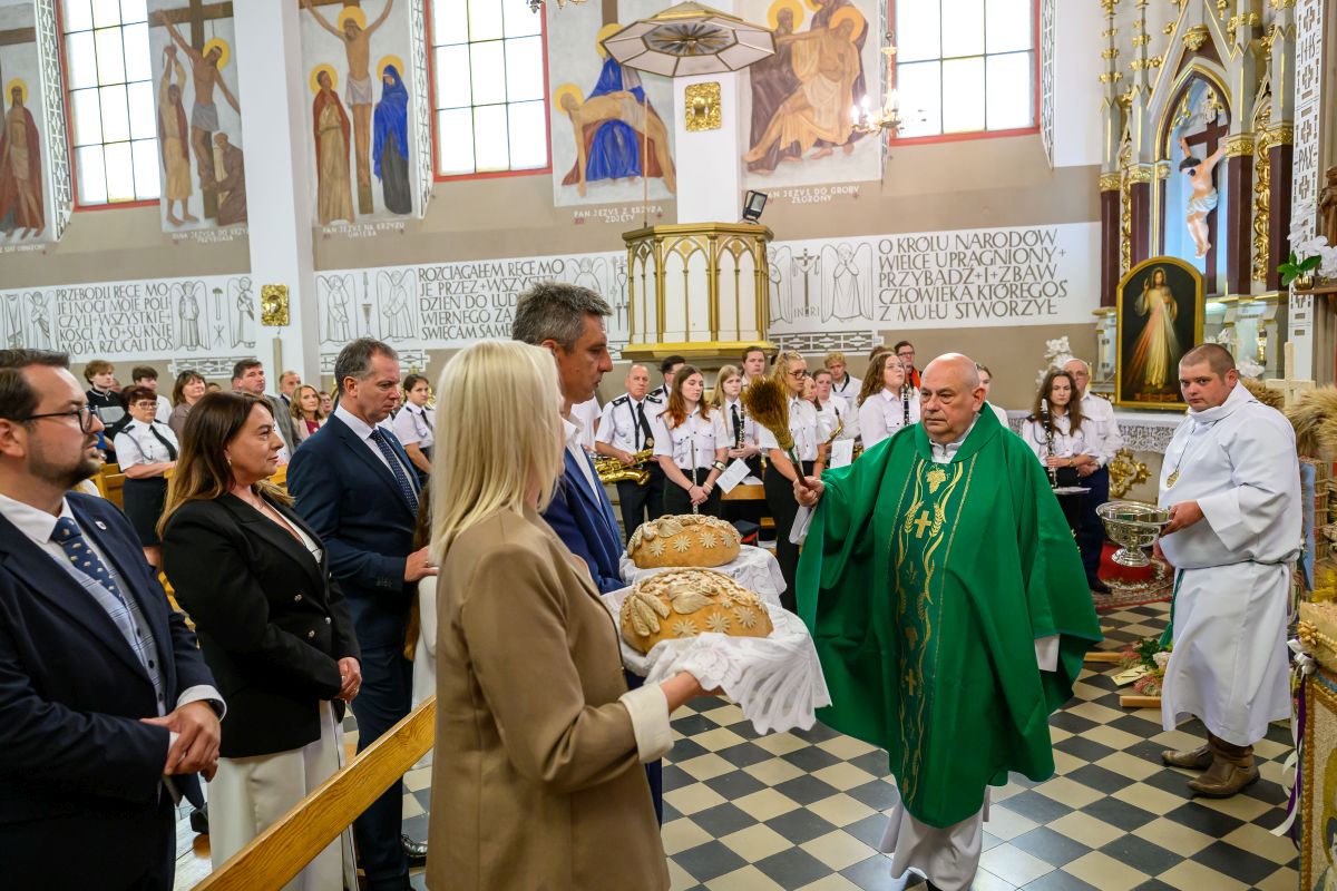 Kapłan w zielonych szatach błogosławi chleb w kościele, otoczony wiernymi i chórem. Wnętrze zdobią freski i witraże.