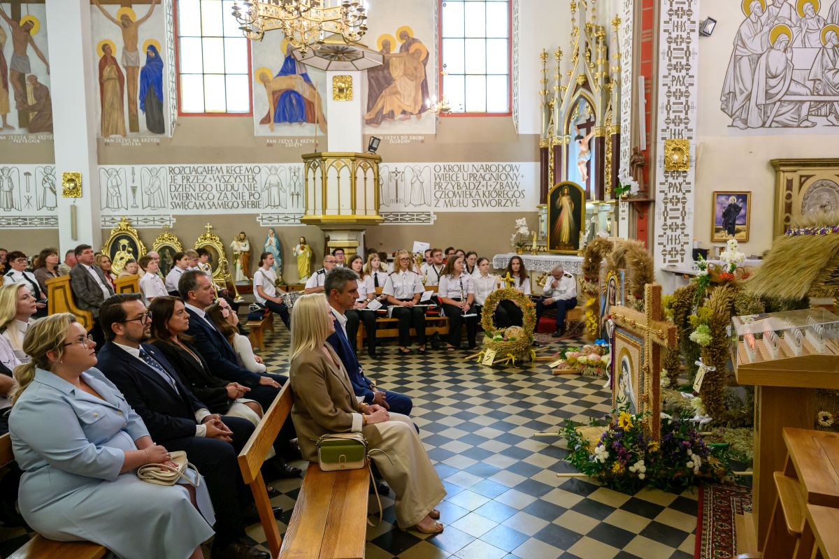 Ludzie w kościele na uroczystości, siedzą na ławkach. Dekoracje z kwiatów i zboża. Tło z freskami i witrażami.