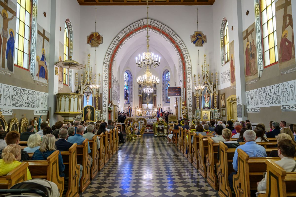 Wnętrze kościoła z tłumem wiernych. Kolorowe witraże i freski zdobią ściany. W centrum widoczny jest bogato zdobiony ołtarz.