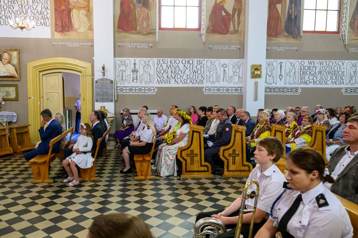 Obraz przedstawia ludzi siedzących w kościele na drewnianych ławkach. W tle są malowidła ścienne i cytaty. Kolorystyka jest jasna, dominują beże i brązy.