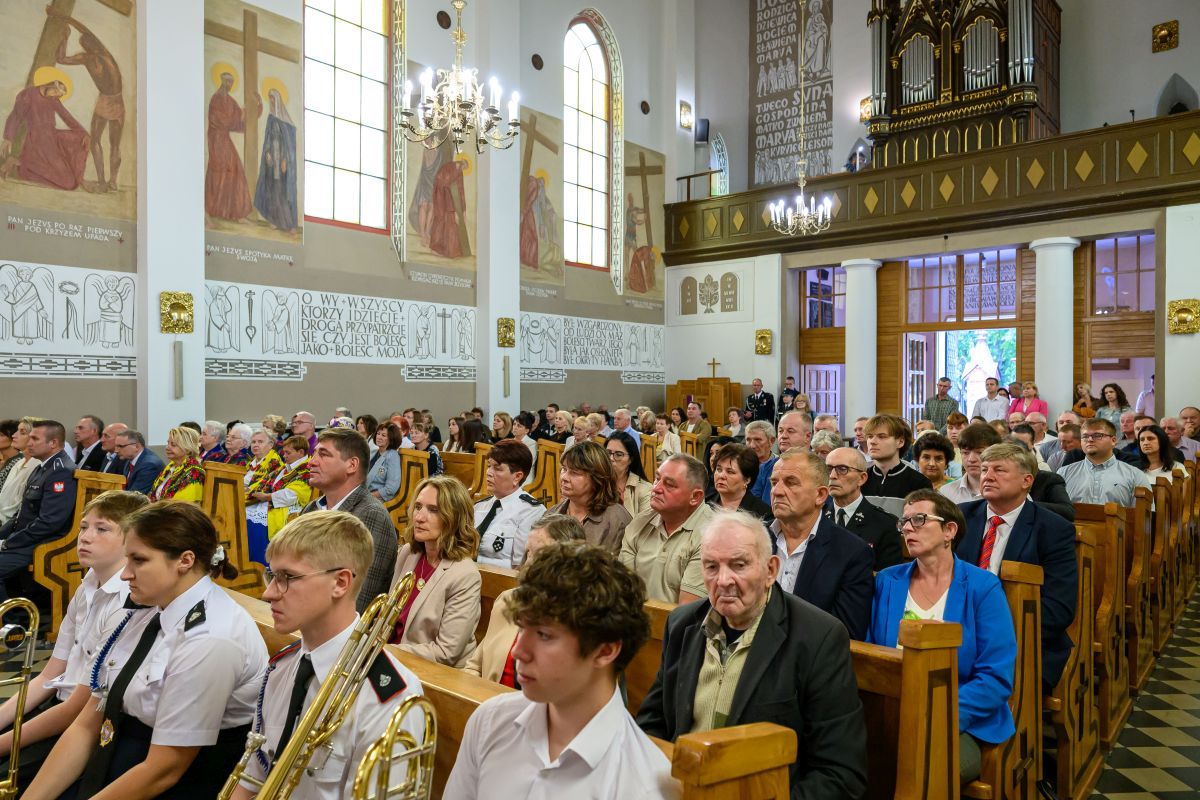 Ludzie siedzą w kościele ozdobionym freskami i witrażami. Jasne wnętrze z drewnianymi ławkami i złotymi elementami. Na ścianach wiszą obrazy religijne.