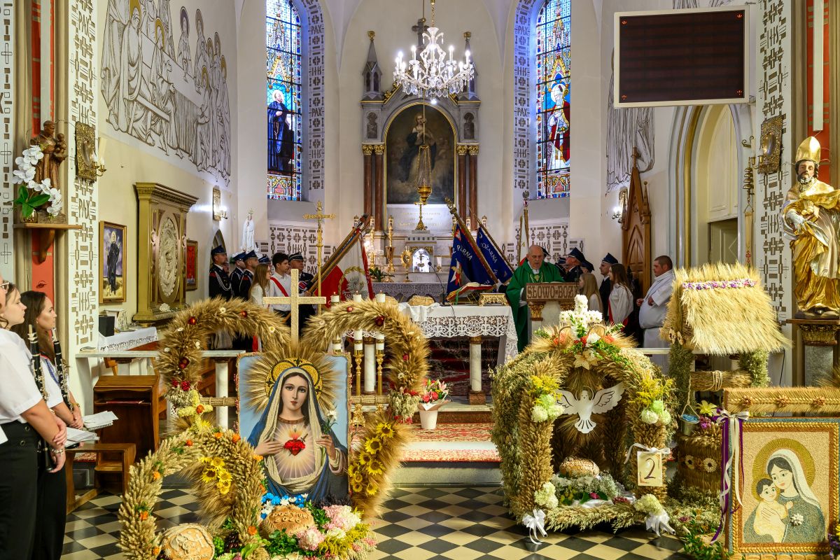 Kościół wewnątrz z ozdobnymi wieńcami zbożowymi i obrazem Maryi. W tle ołtarz, witraże, ksiądz przy ambonie. Kolory jasne, złote i zielone.
