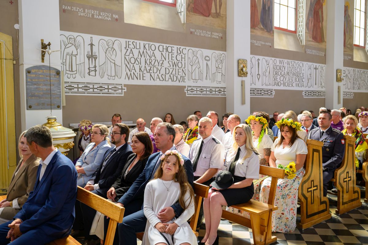 Wnętrze kościoła wypełnione ludźmi w eleganckich ubraniach. Na ścianach są malowidła i napisy religijne. Kilka osób ma wianki z kwiatów.