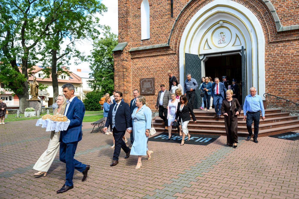 Ludzie wychodzą z kościoła z czerwonej cegły na tle zieleni i nieba. Osoby są elegancko ubrane. Mężczyzna niesie chleb na białej serwetce.