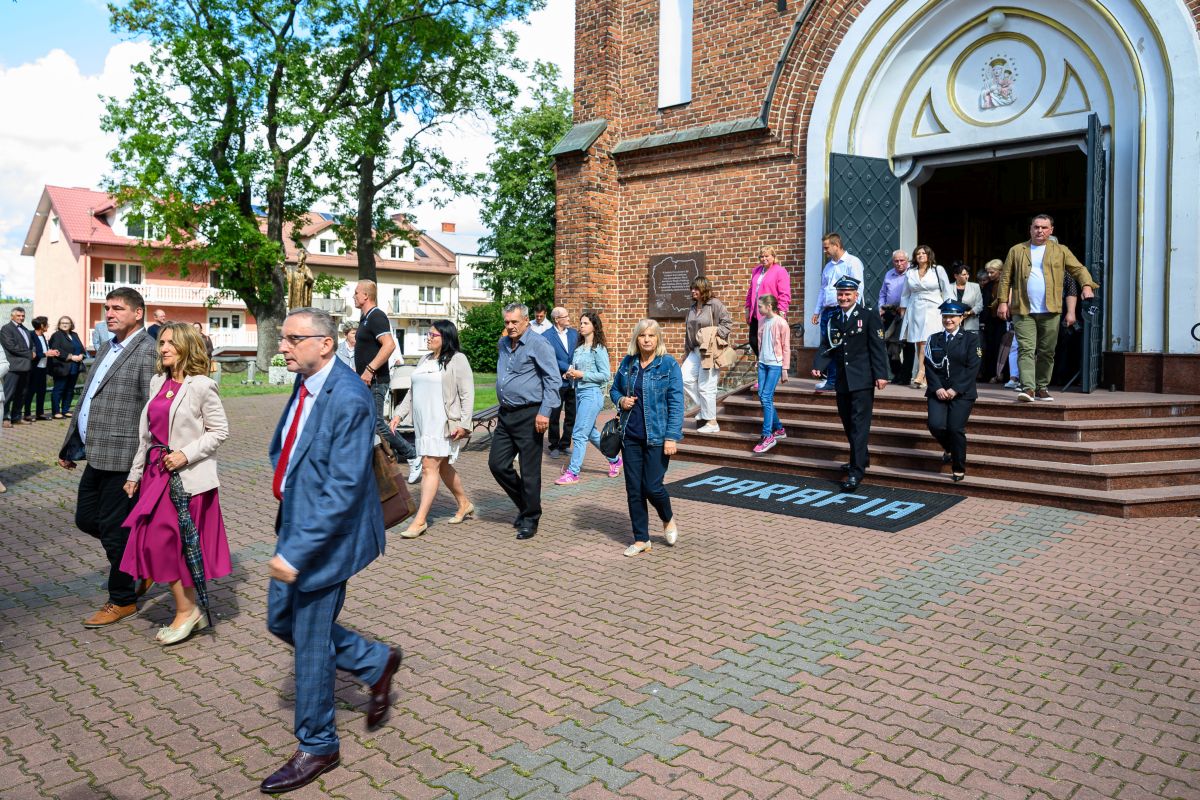 Grupa ludzi wychodzi z kościoła z ceglaną fasadą. Tło to drzewa i budynki. Kolory są jasne, z granatowymi i różowymi akcentami.