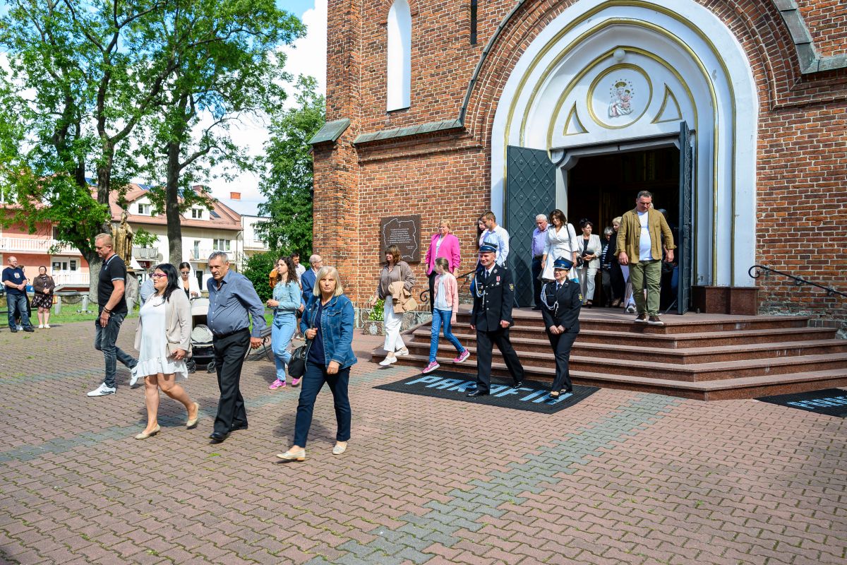 Ludzie wychodzą z ceglanego kościoła o białym wejściu. Na dziedzińcu ukazują się drzewa i budynki. Panuje słoneczny dzień.