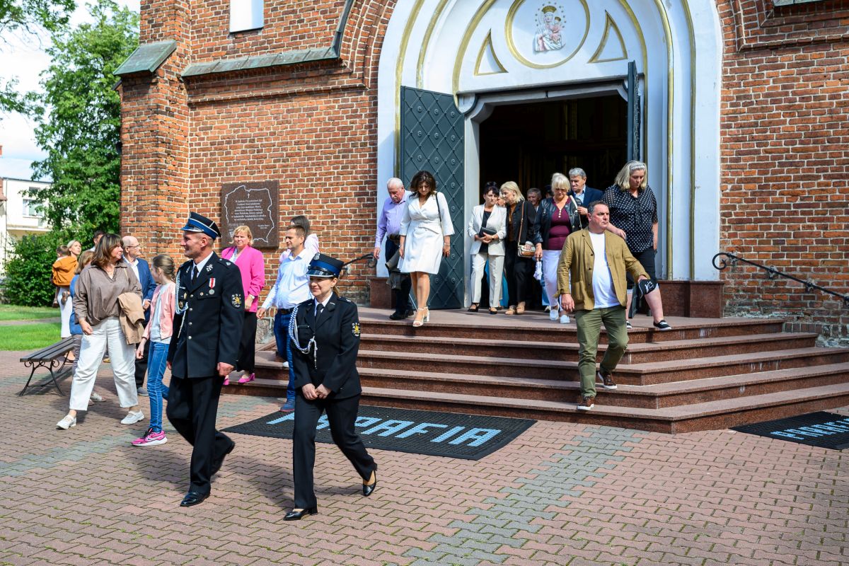 Grupa ludzi wychodzi z ceglanego kościoła z białym wykończeniem, na schodach widoczni strażacy. Słoneczny dzień z zielenią w tle.