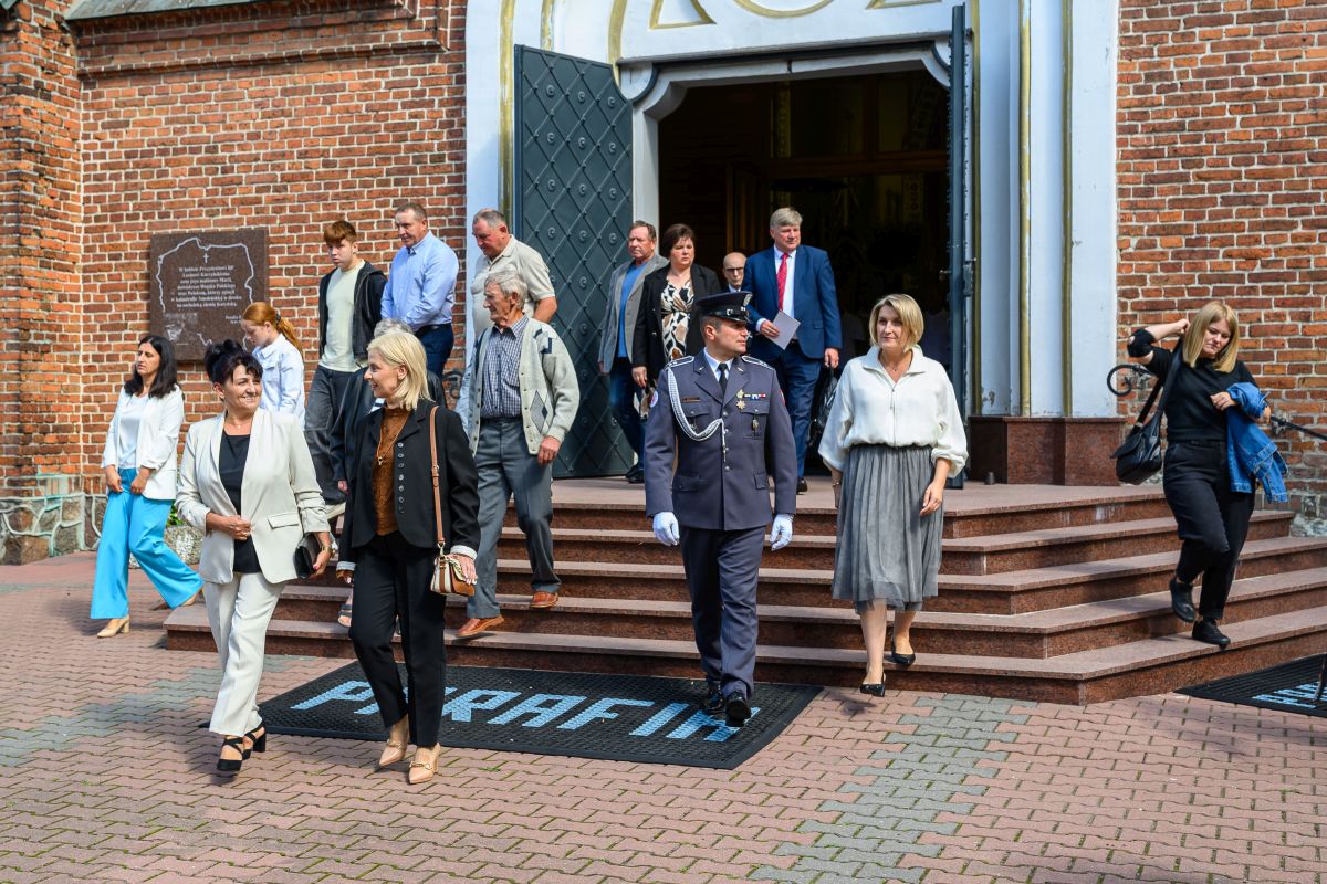 Grupa ludzi opuszcza ceglaną budowlę z dużymi drzwiami. Ubrani są elegancko, widać mundur. Tło to czerwona cegła i schody.