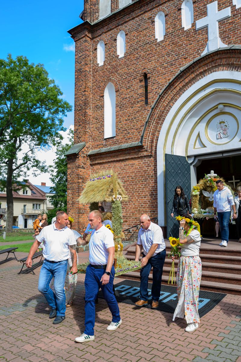 Przed kościołem z cegły, ludzie w białych koszulach niosą ozdobne konstrukcje ze słomy. Tło to błękitne niebo i drzewo. Obok drzwi stoi kobieta z wiankiem z kwiatów.