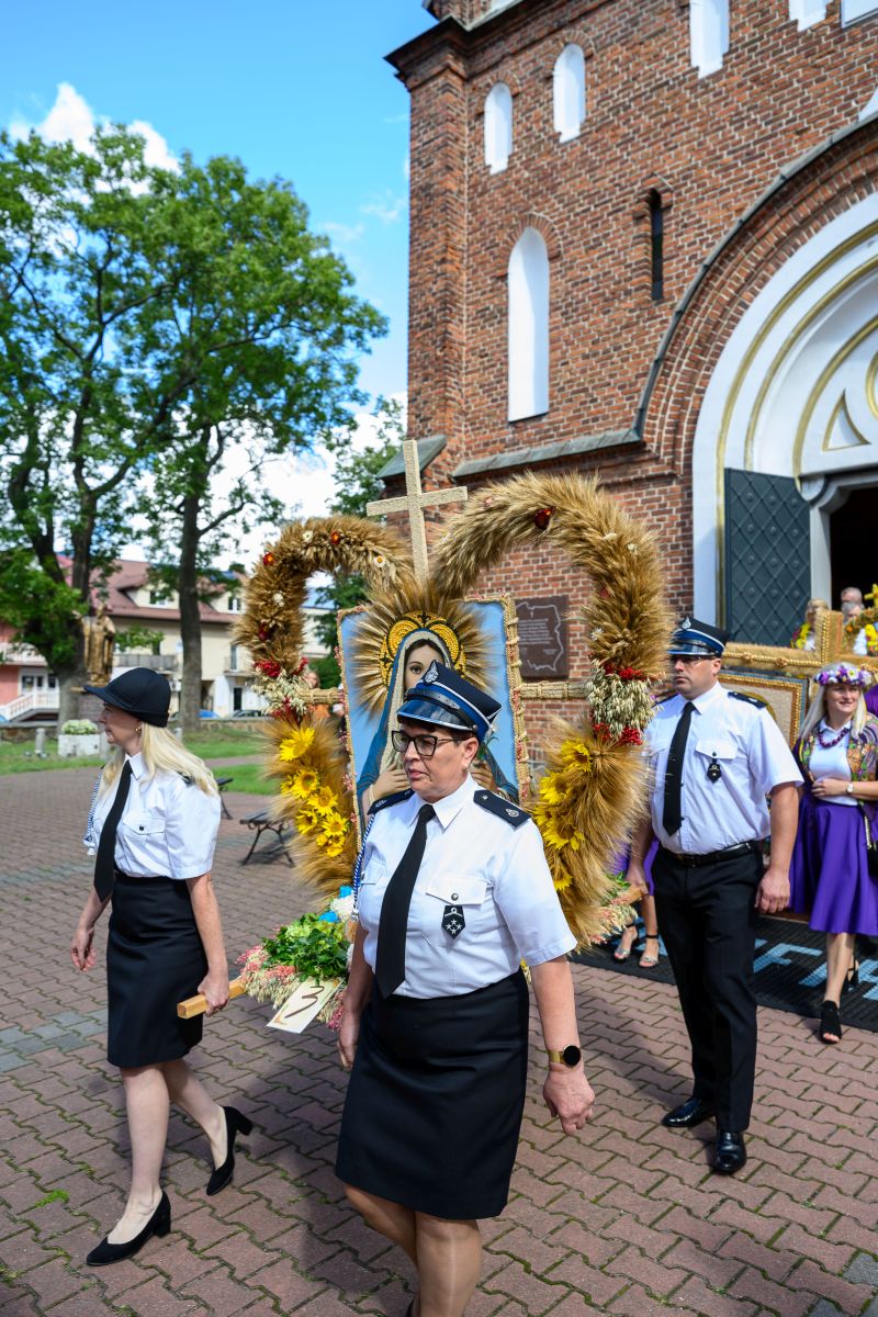 Ludzie w mundurach niosą wieniec z wizerunkiem Matki Boskiej przed ceglaną świątynią. Zieleń drzew i niebieskie niebo wokół. Dominują kolory bieli, złota i brązu.
