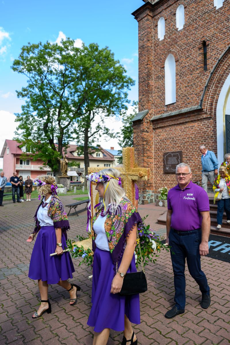 Ludzie w fioletowych strojach tradycyjnych przed ceglanym kościołem. Jasne niebo, zielone drzewo w tle. Korzystają z uroczystości.
