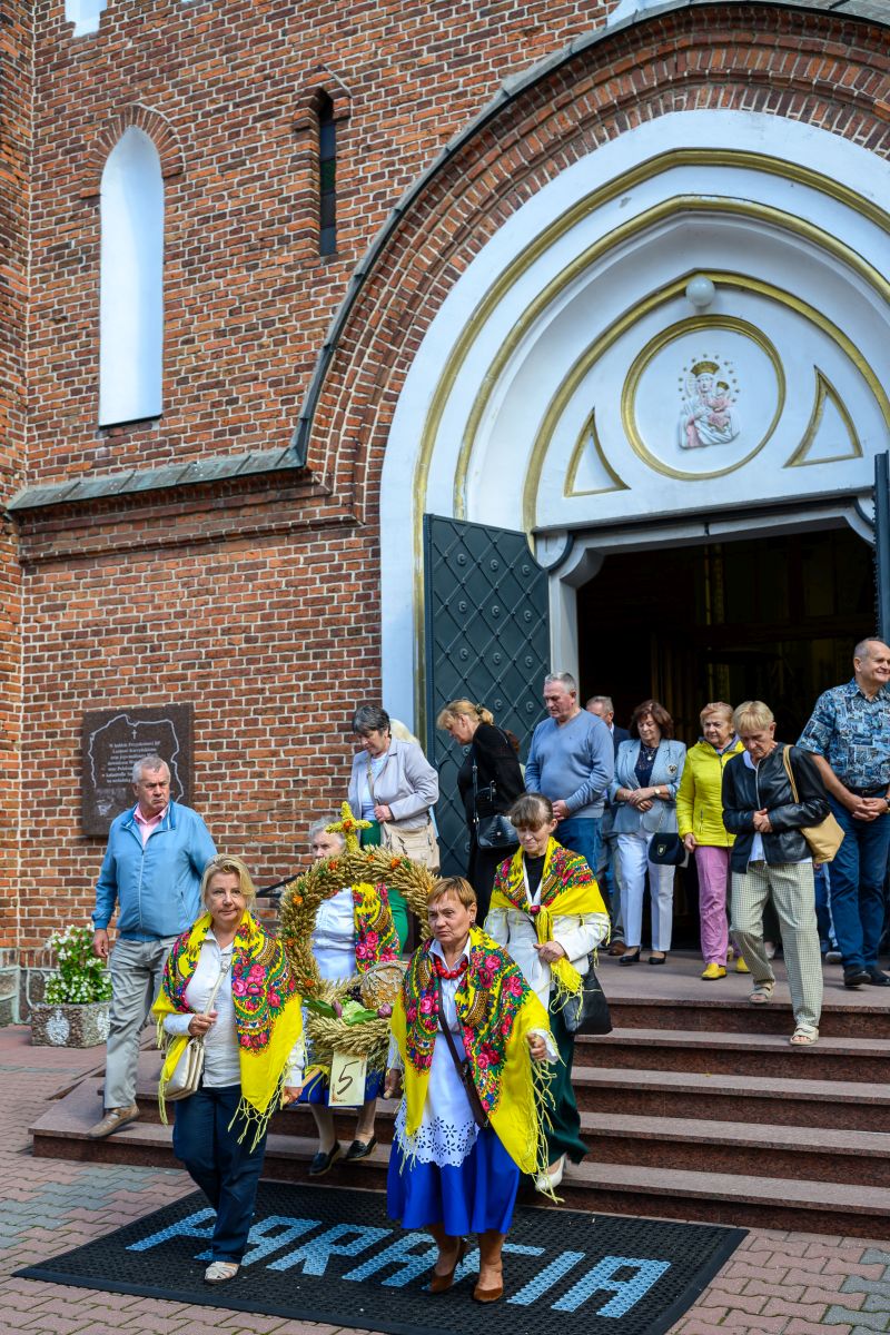 Grupa ludzi opuszcza ceglaną świątynię z ozdobną bramą. Kobiety w kolorowych strojach niosą żniwne wianki. Tło z cegły i białymi detalami.