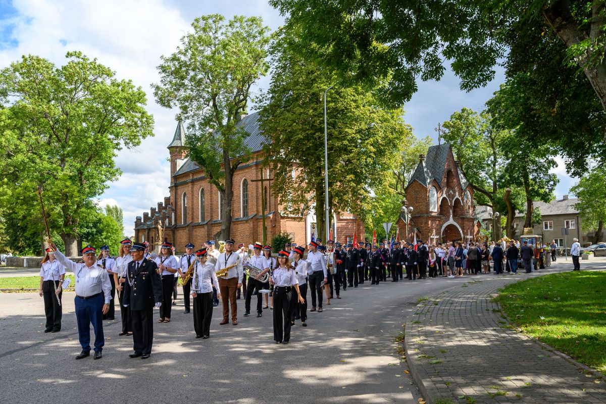 Parada ludzi w mundurach maszerujących wzdłuż ceglanego kościoła, zielone drzewa, jasne niebo.